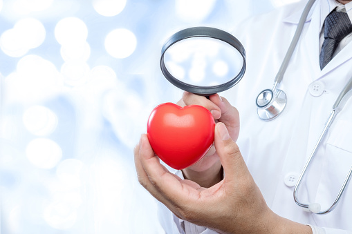 Professional medical doctor holding a magnifying glass check up on a red heart ball on blur office in the hospital and bokeh background. Concept of health care.