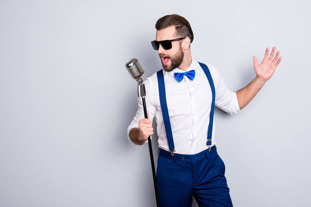 Portrait of handsome attractive singer in blue pants with suspenders and black glasses, singing hit with open mouth in microphone gesture with hand isolated on grey background